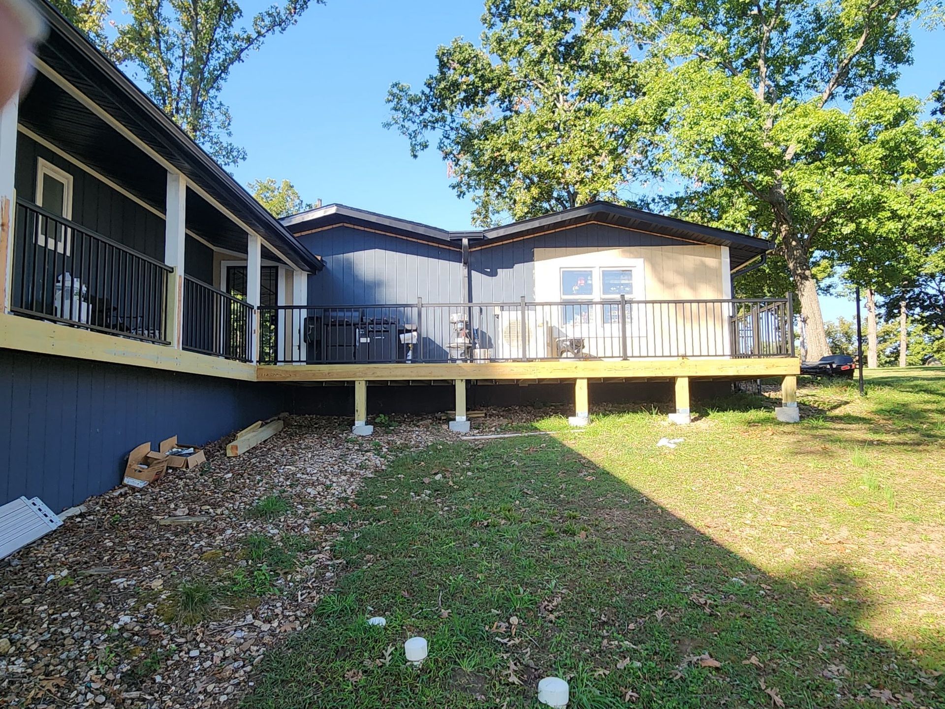 Deck extension with black railing attached to a blue house with trees in the background on a sunny day.