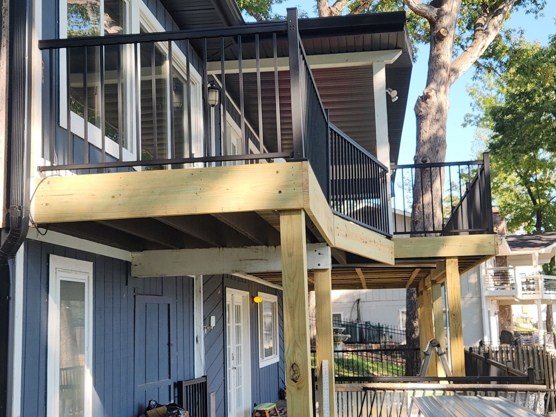 Wooden deck with black railing attached to a blue house with windows. A tree is growing through the deck.