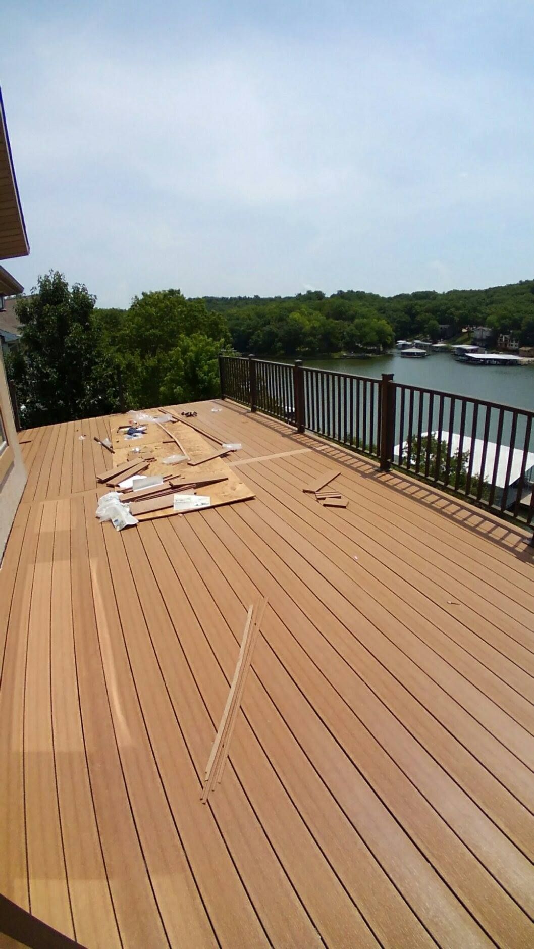 Wooden deck overlooking a lake, with supplies scattered. Black railing and trees in background.