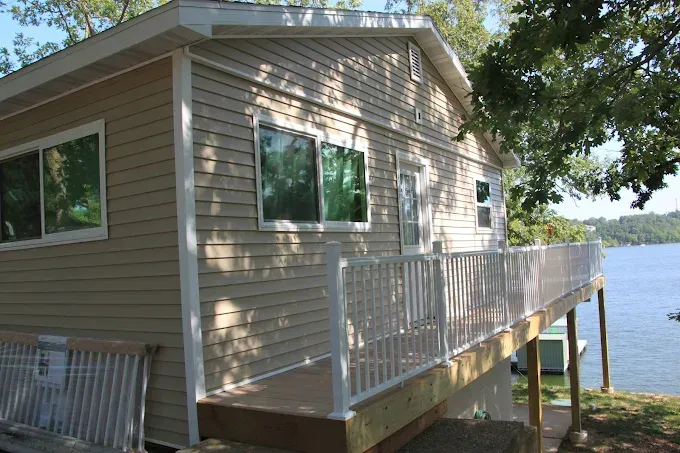 Tan sided house with white trim, deck overlooking a lake.