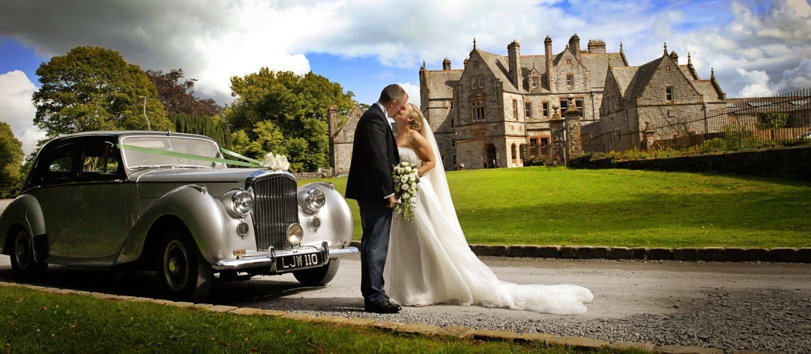A bride and groom are kissing in front of a silver car.