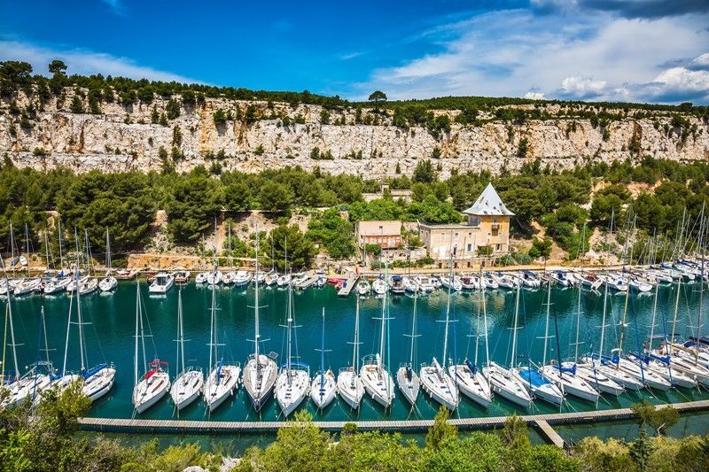A row of boats are docked in a harbor with a cliff in the background.