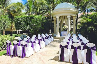 A row of white chairs with purple bows are lined up in front of a gazebo.