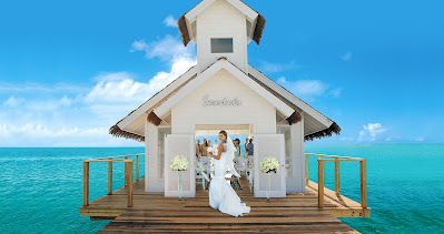 A bride and groom are standing in front of a small white church overlooking the ocean.
