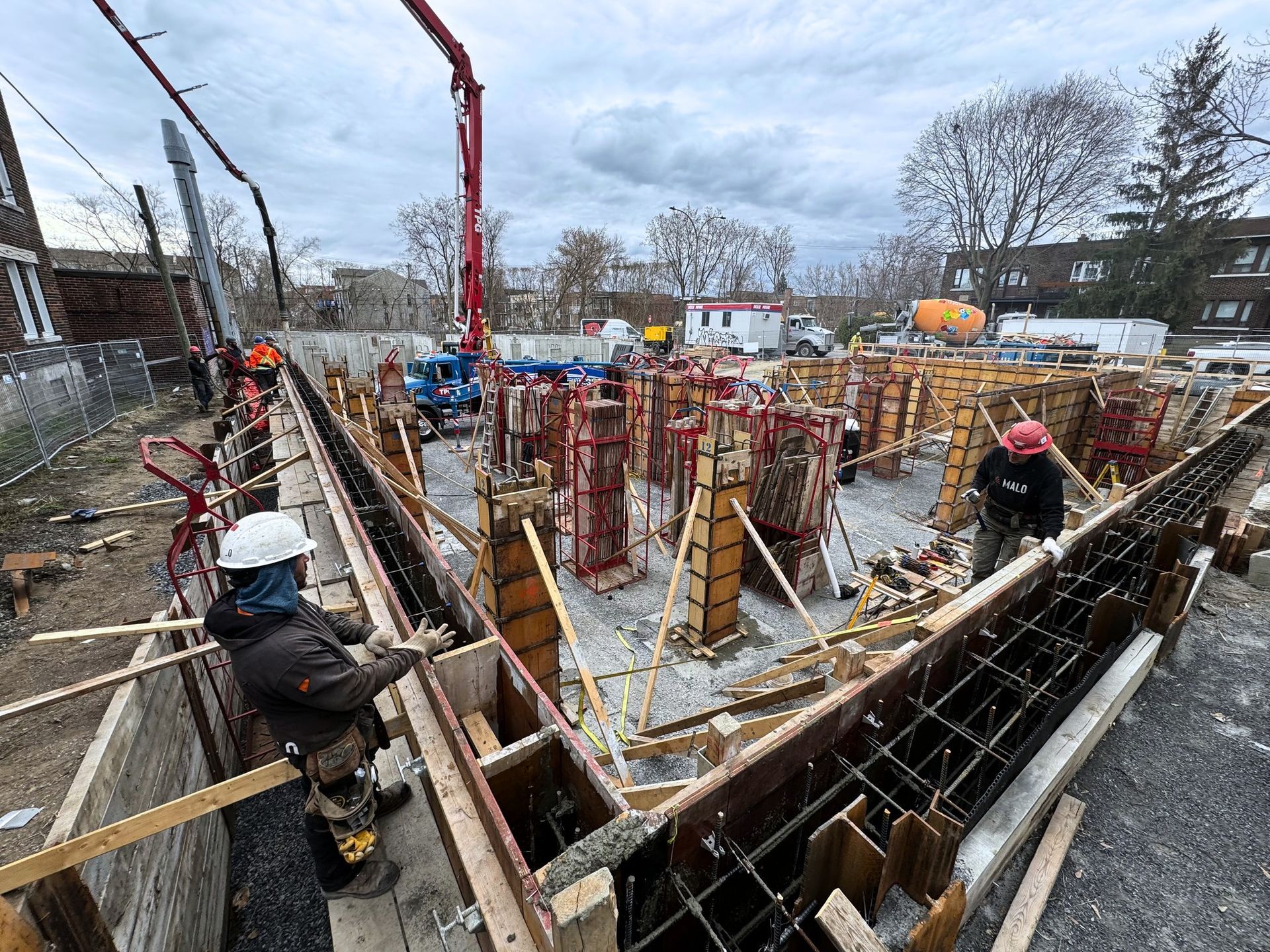 Des ouvriers du bâtiment coulent du béton sur un chantier de fondations à l'aide d'un camion-pompe à béton, sous un ciel nuageux.