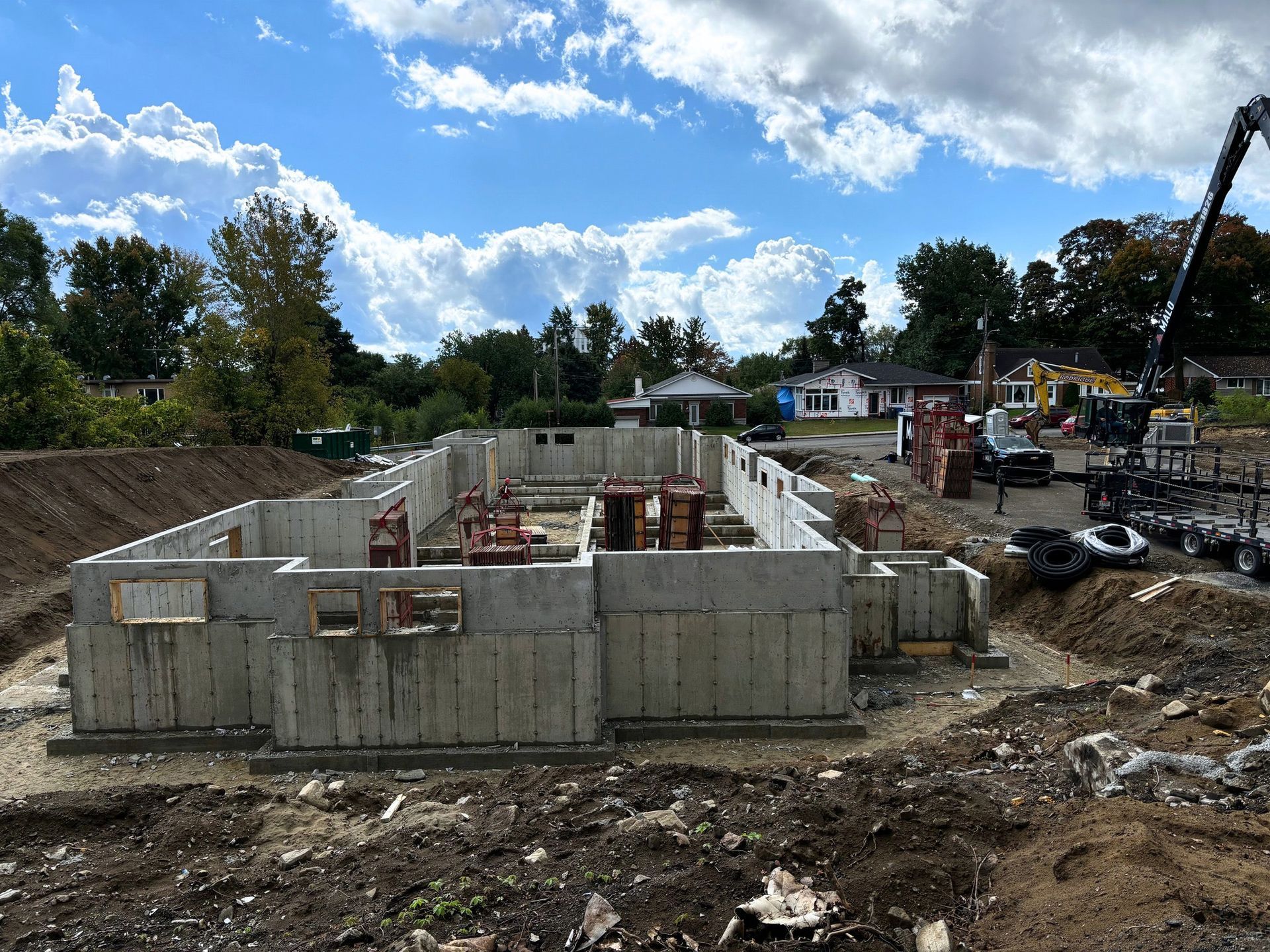 Fondations d'un bâtiment en construction sur un terrain vague, avec des équipements et d'autres bâtiments visibles en arrière-plan.
