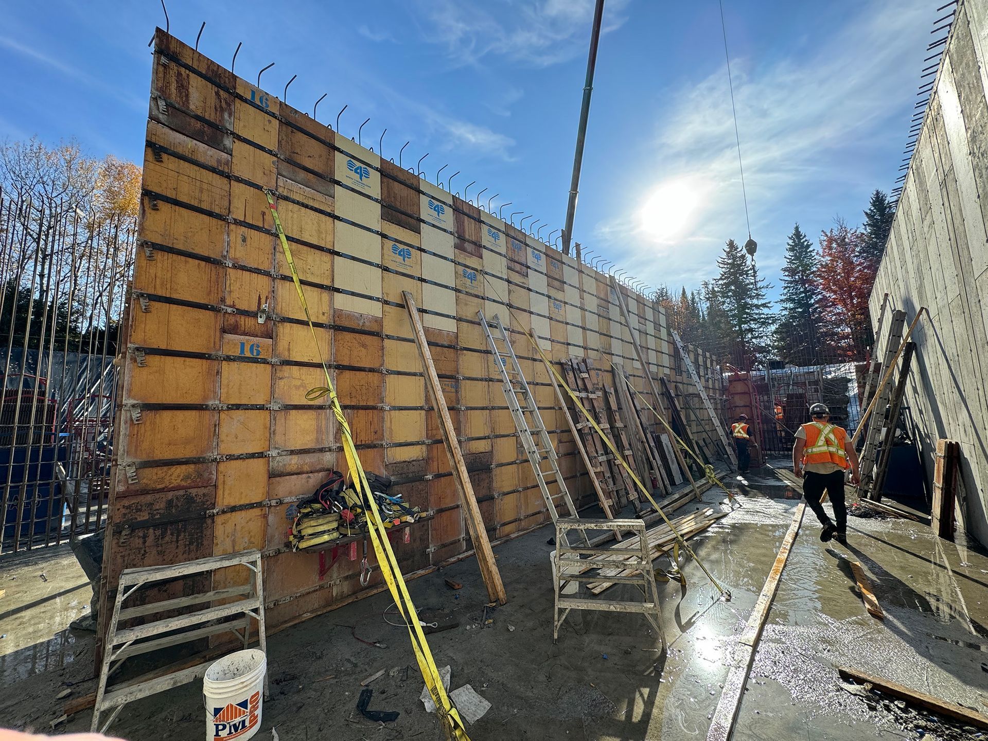 Chantier de construction avec un haut coffrage en bois pour un mur en béton. Un ouvrier en gilet orange passe.