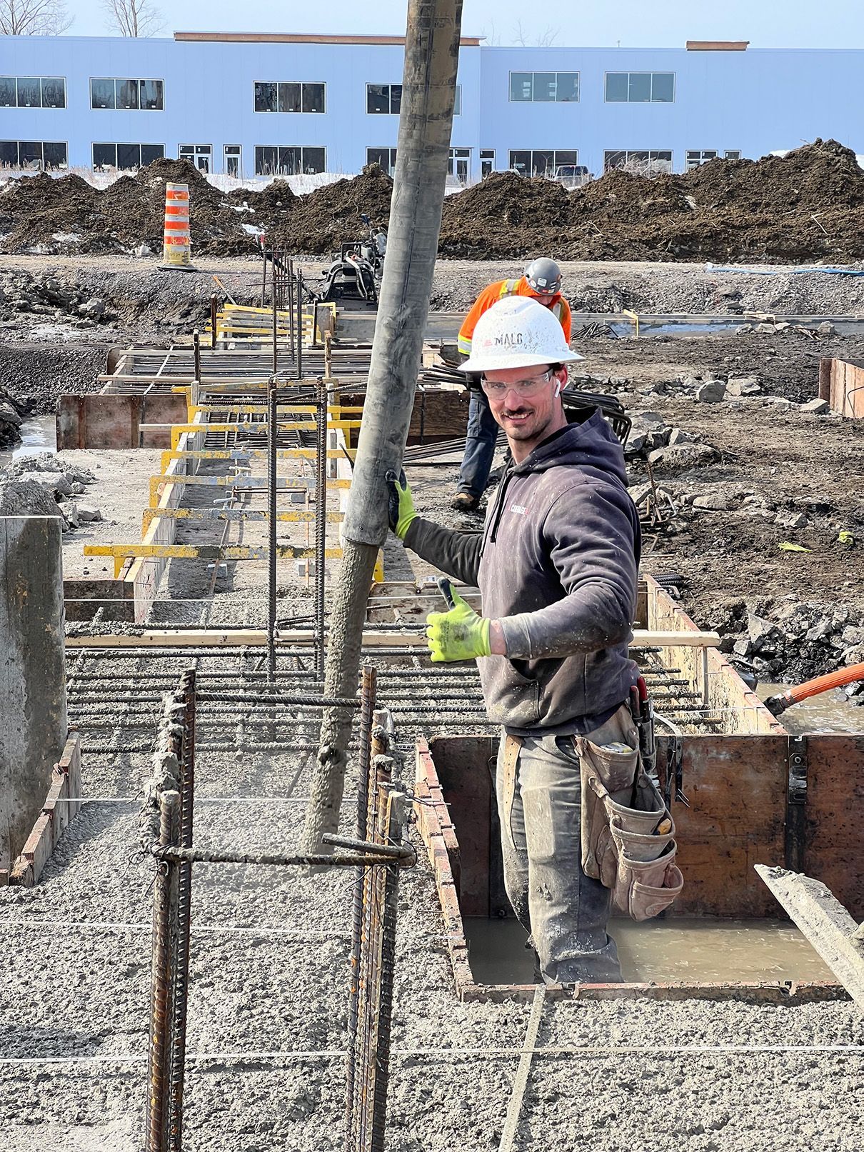 Un ouvrier du bâtiment coule du béton dans un coffrage de fondation à l'aide d'un tuyau d'arrosage. Il porte un casque, des gants et une tenue de travail.