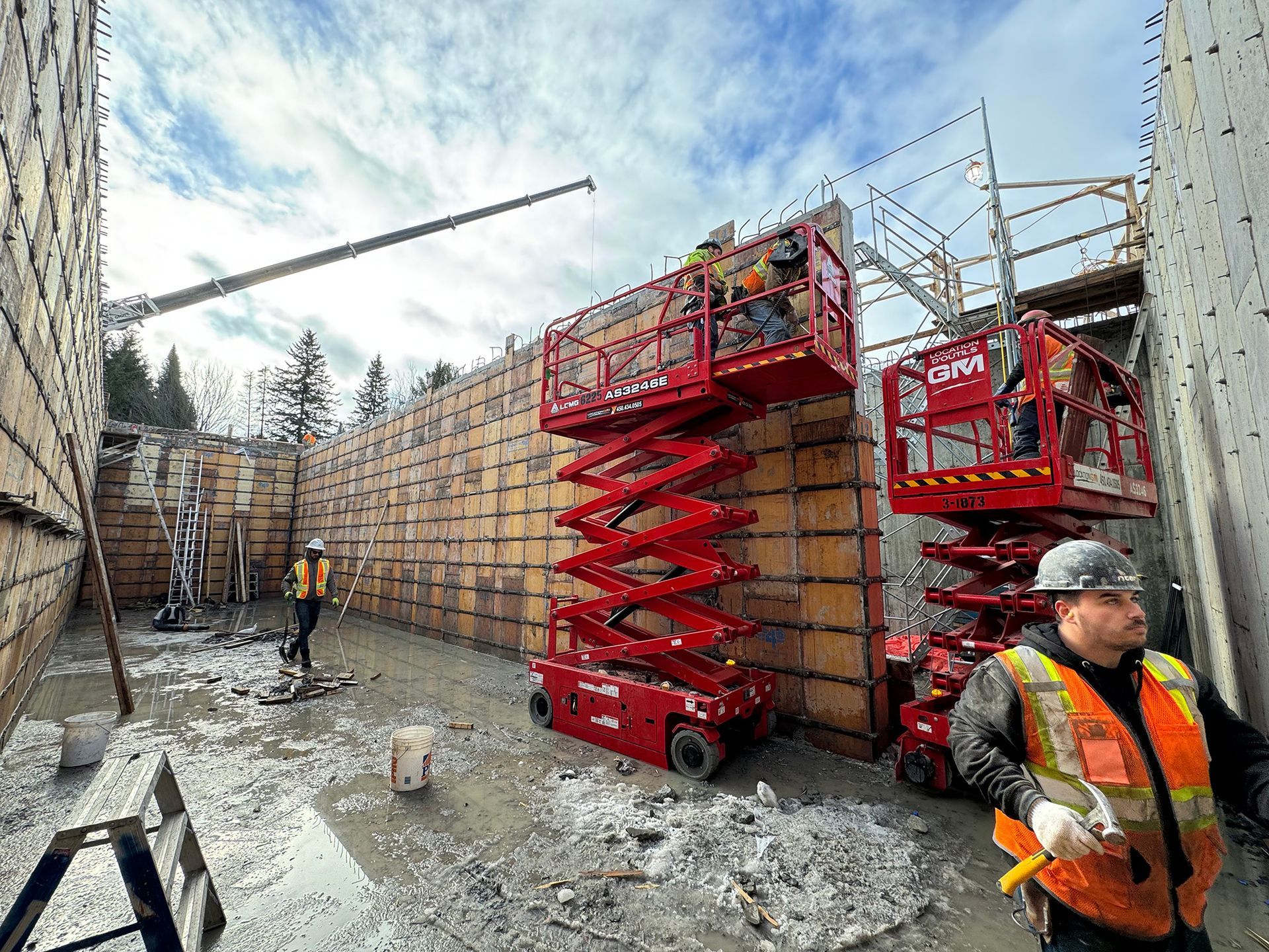 Chantier avec ouvriers, échafaudages et nacelles élévatrices rouges. Des coffrages en béton sont en cours de montage.