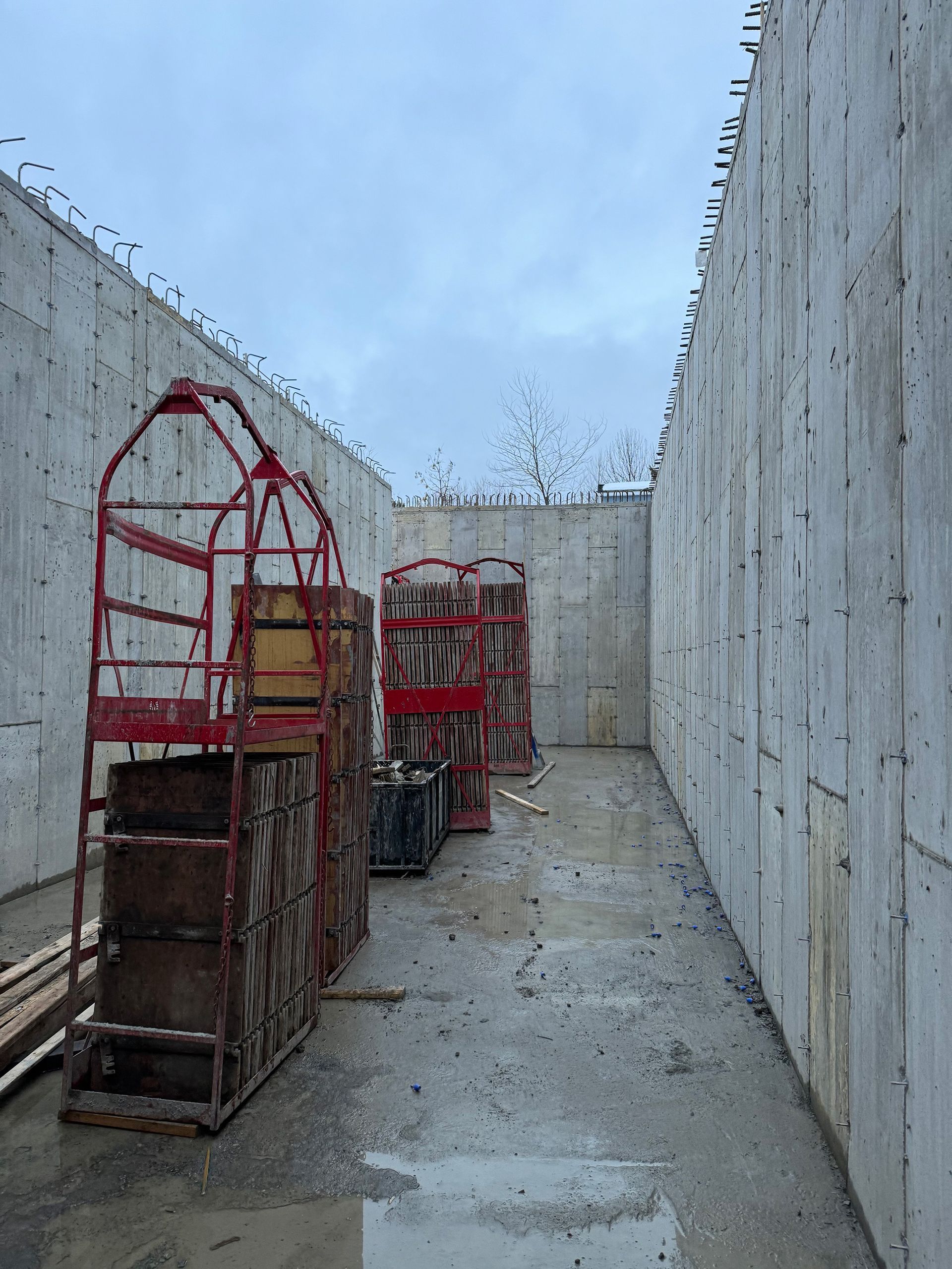 Des murs en béton enserrent un chantier rectangulaire. À l'intérieur, on aperçoit des échafaudages rouges et des coffrages en bois. Ciel couvert.