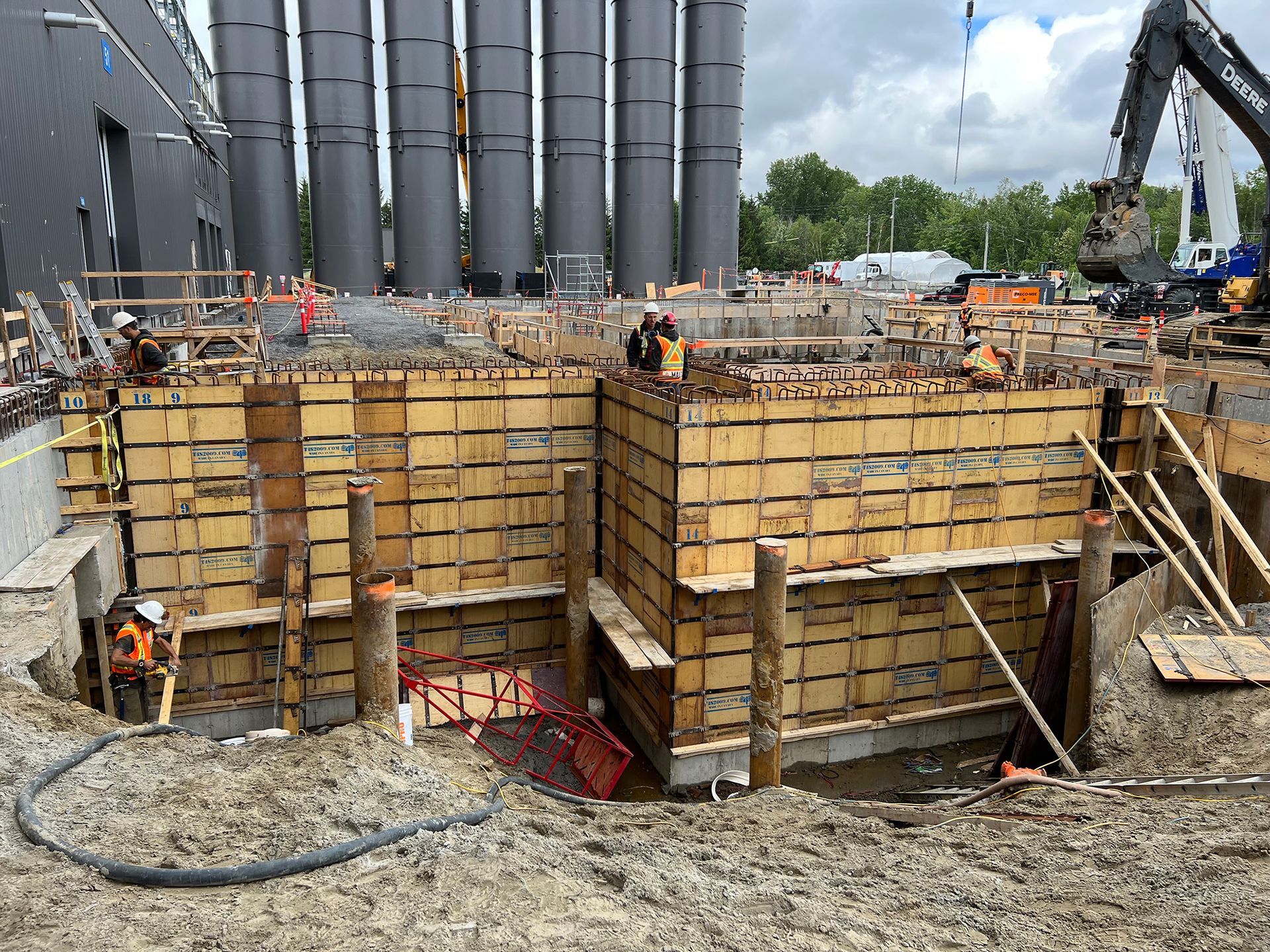 Chantier de construction avec coffrage en bois pour murs en béton ; ouvriers, excavatrice et grands réservoirs en arrière-plan.