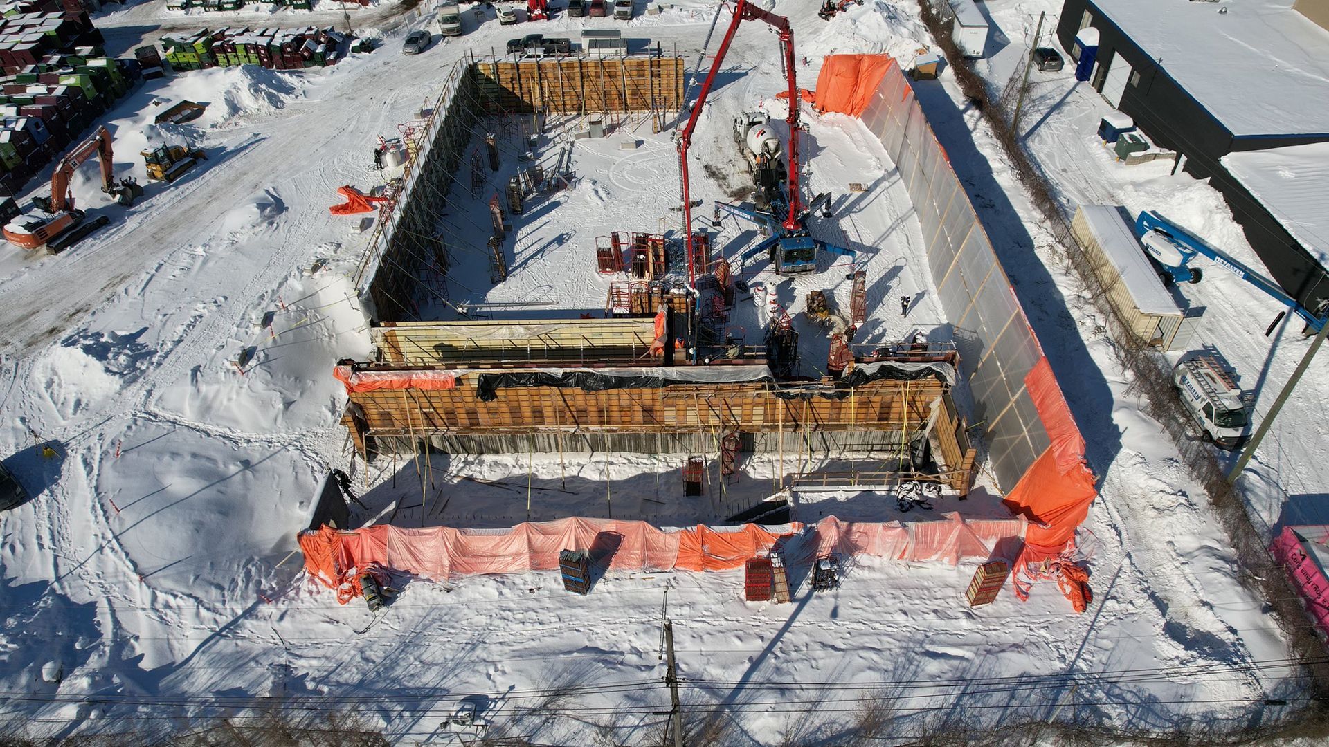 Chantier de construction sous la neige ; coulage de béton dans une fosse de fondation rectangulaire protégée par une barrière orange.