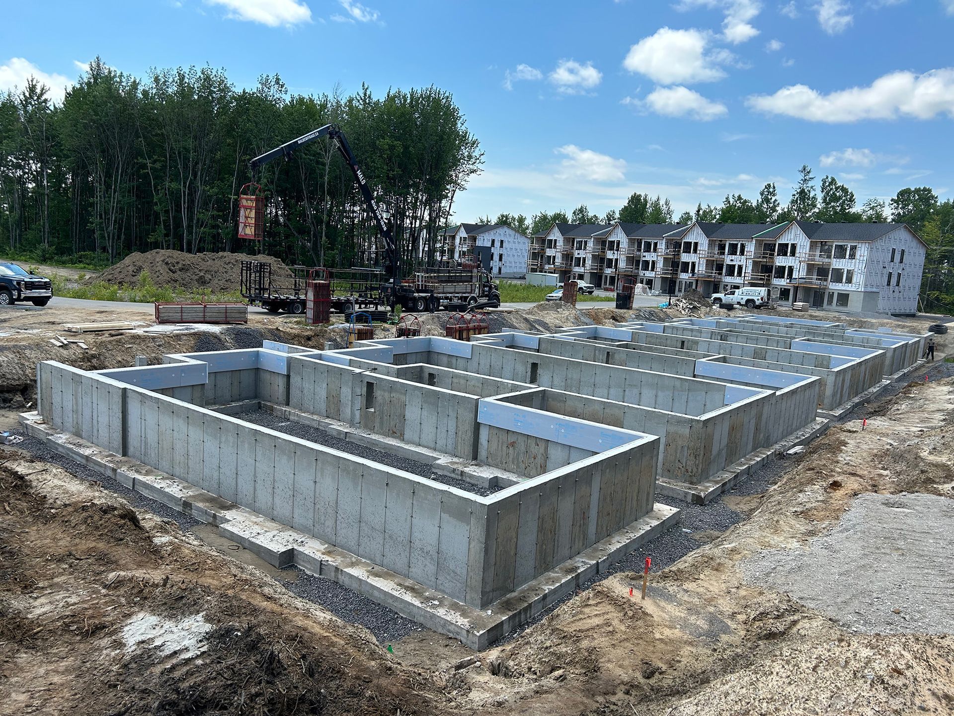 Chantier de construction avec fondations en béton. On aperçoit une grue, de la terre et des maisons de ville en construction.