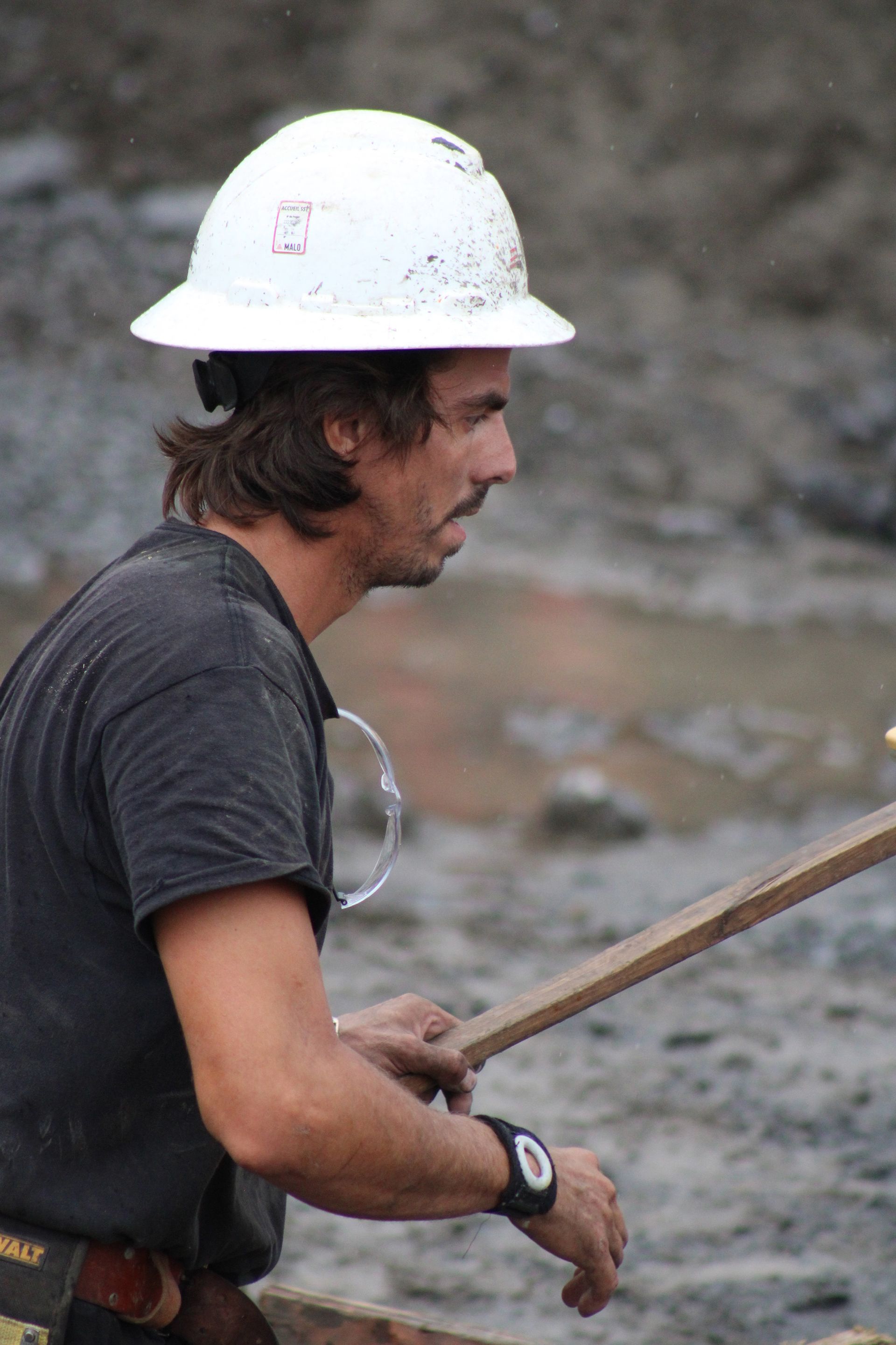 Ouvrier du bâtiment portant un casque blanc, tenant une poutre en bois à l'extérieur.