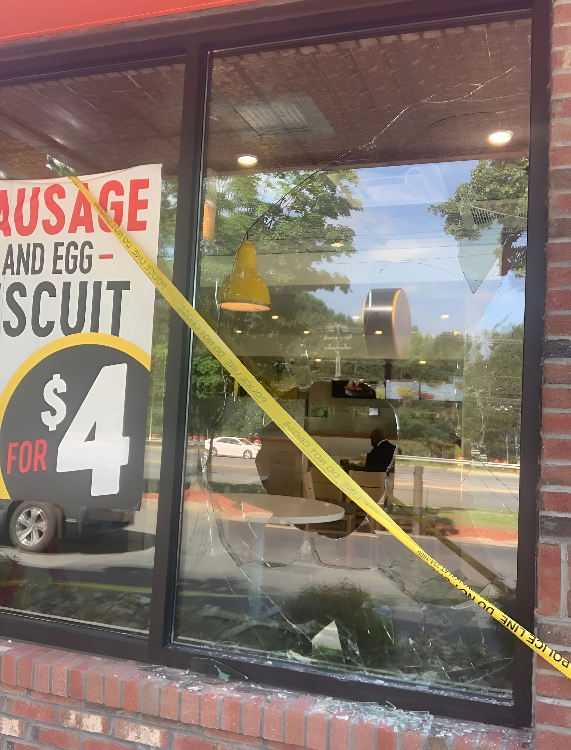 Shattered window of a fast food restaurant with yellow caution tape across it. A sign advertises a breakfast biscuit.