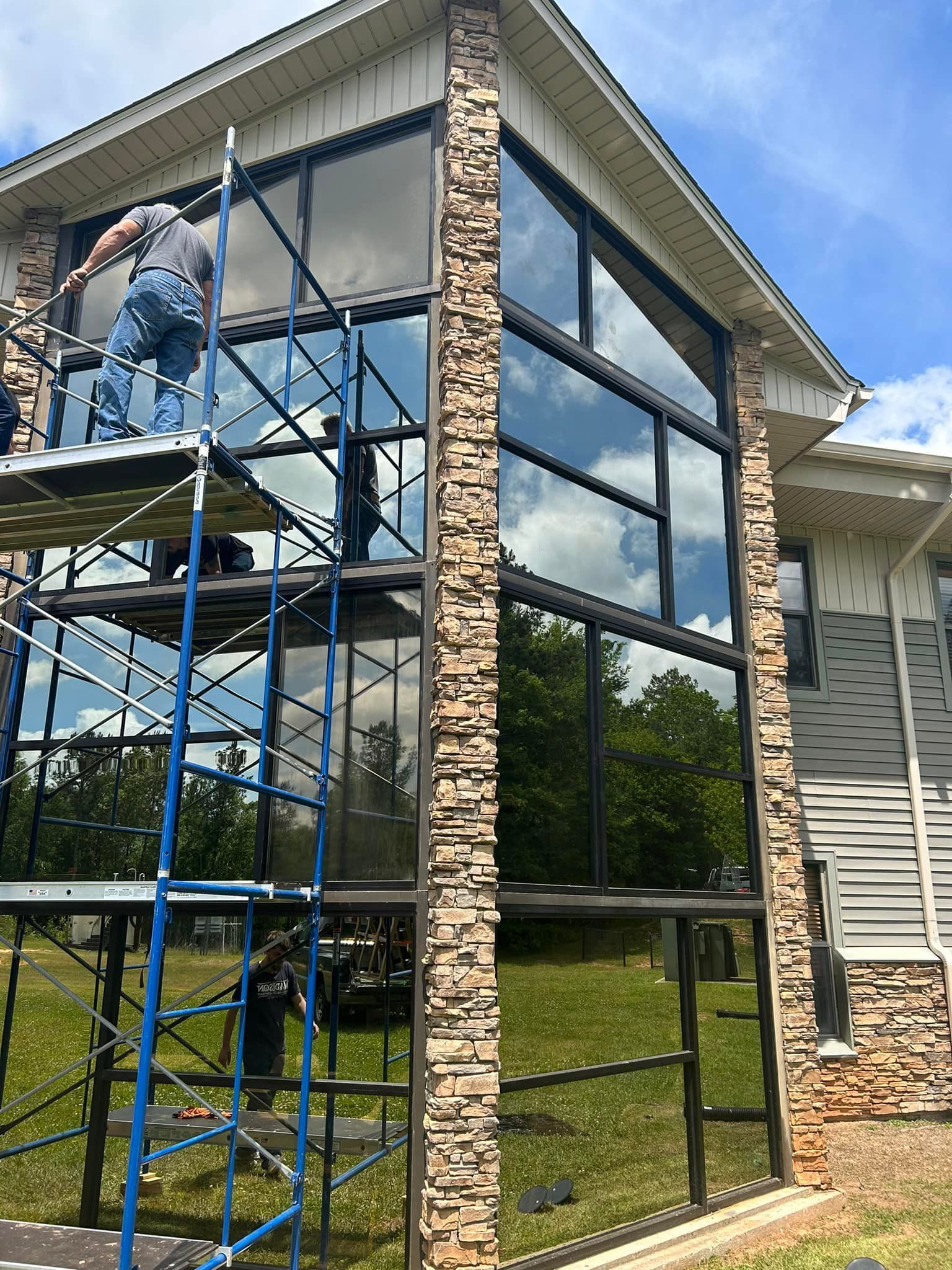 Workers on scaffolding installing large, reflective windows on a house with stone accents.