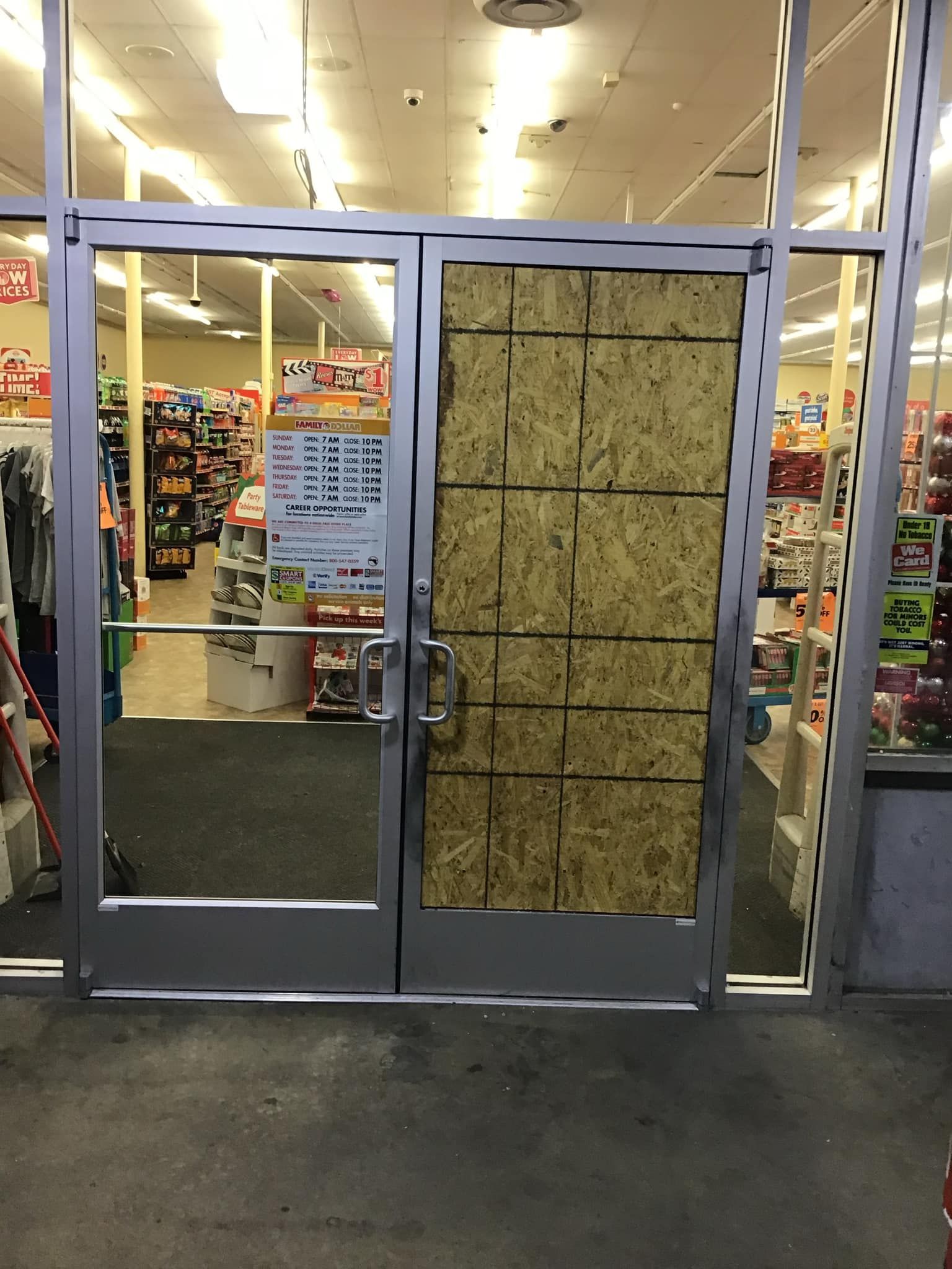 Boarded-up glass doors of a store, possibly due to damage. Inside, merchandise is visible.