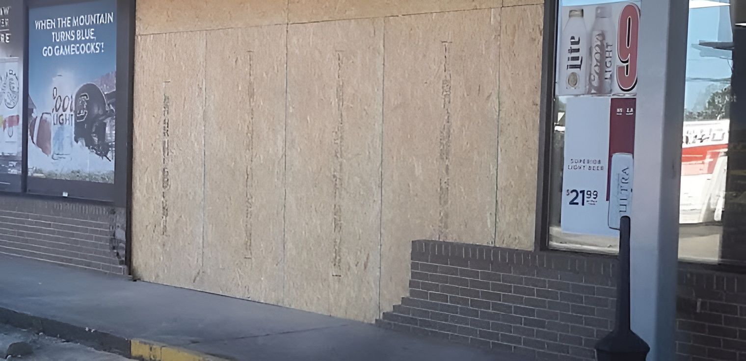 Exterior of a store front with boarded-up windows, brick and concrete, and signage visible.