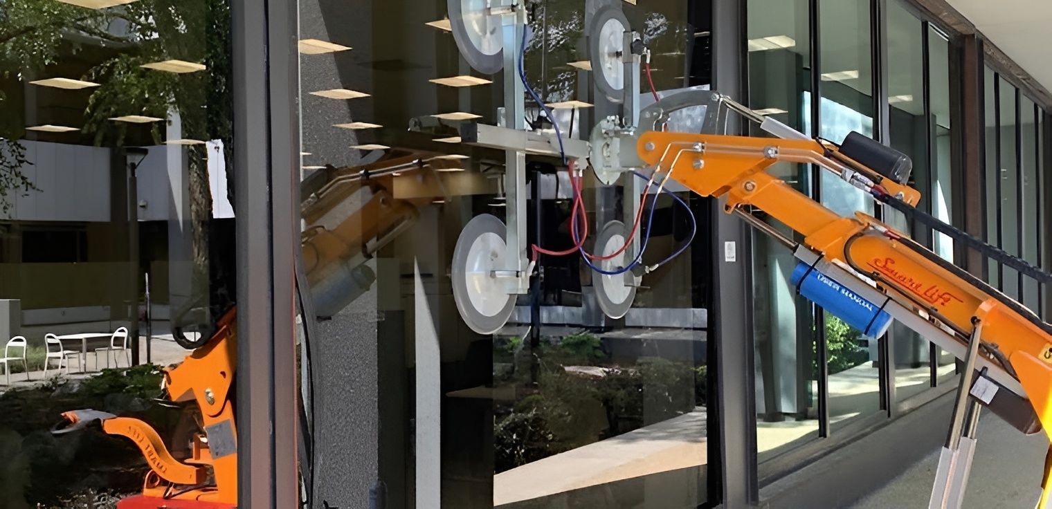 An orange robotic arm with suction cups lifting a glass window on a building.