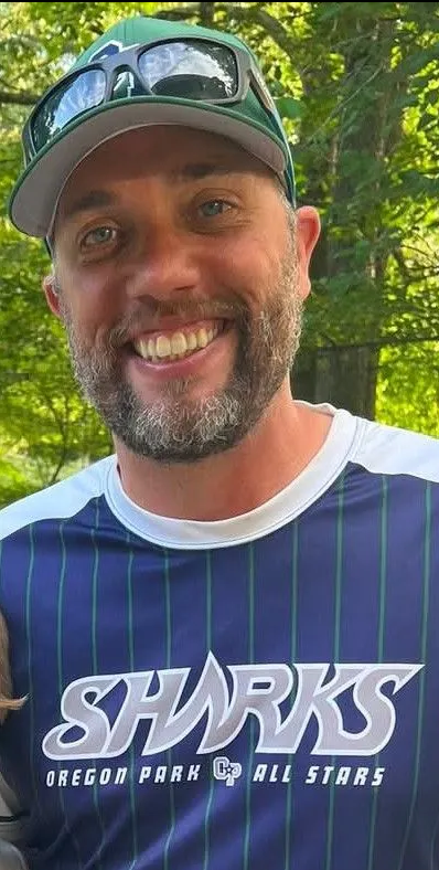 Football coach with arms crossed, looking stern on a sunny field, players in the background.