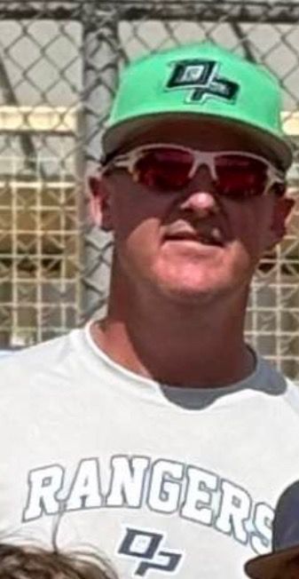 Football coach with arms crossed, standing on a football field. He wears a navy polo shirt and gray pants.