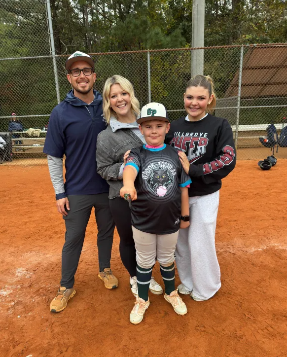 Football coach with arms crossed, standing on a football field. He wears a navy polo shirt and gray pants.