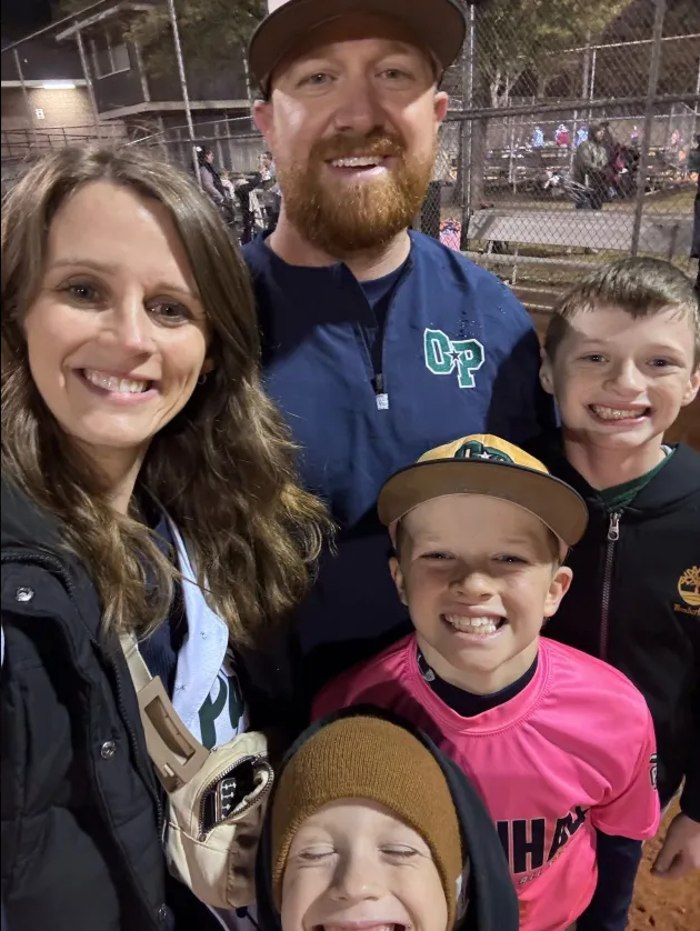 Football coach with arms crossed, looking stern on a sunny field, players in the background.