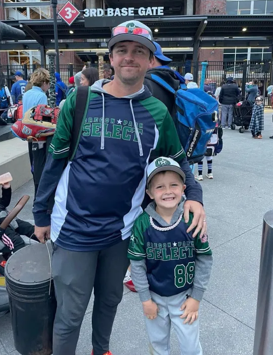 Football coach with arms crossed, standing on a football field. He wears a navy polo shirt and gray pants.