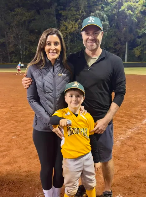 Football coach with arms crossed, looking stern on a sunny field, players in the background.