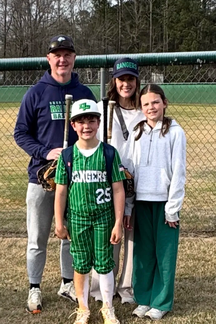 Football coach with arms crossed, looking stern on a sunny field, players in the background.