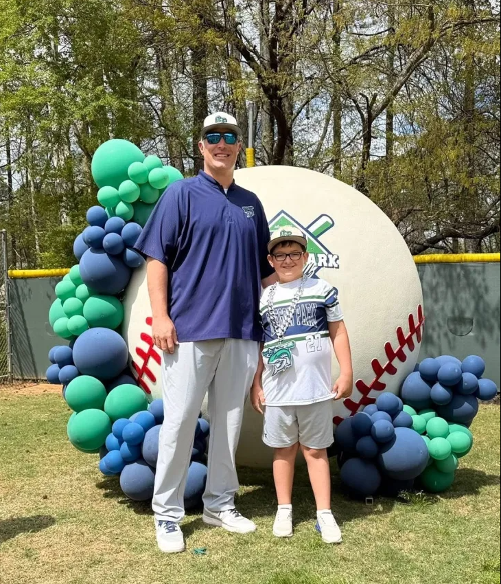 Man in green polo stands on a football field, arms crossed, stadium in background.