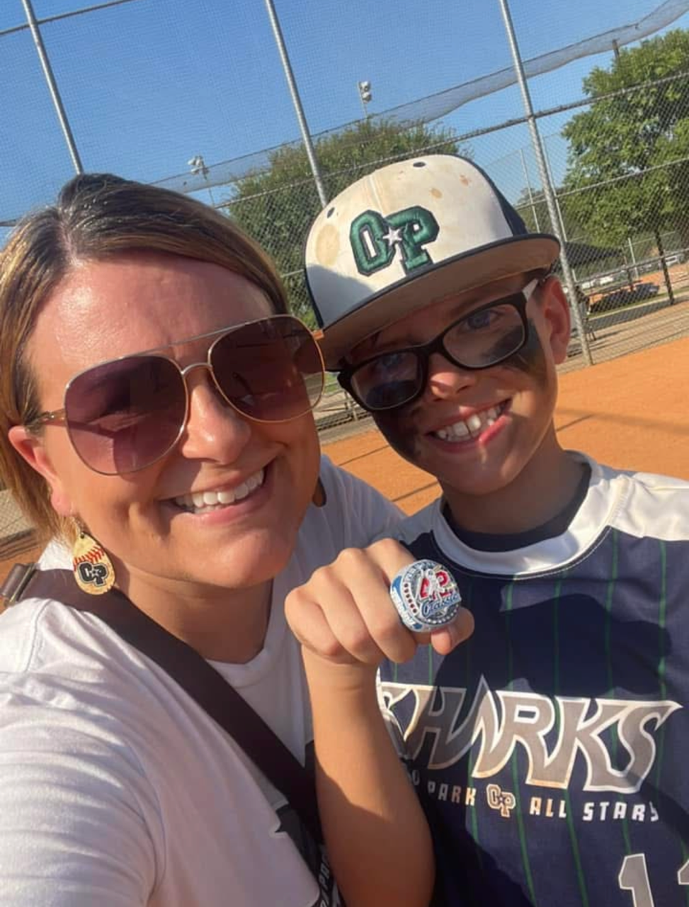 Football coach with arms crossed, looking stern on a sunny field, players in the background.