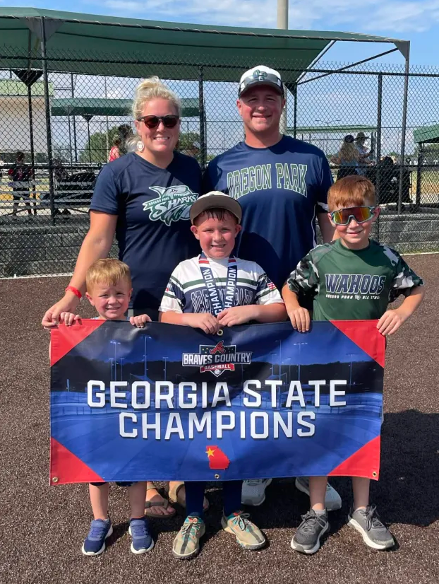 Football coach with arms crossed, standing on a football field. He wears a navy polo shirt and gray pants.