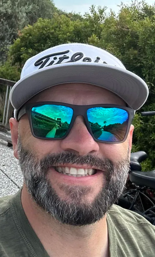 Football coach with arms crossed, looking stern on a sunny field, players in the background.