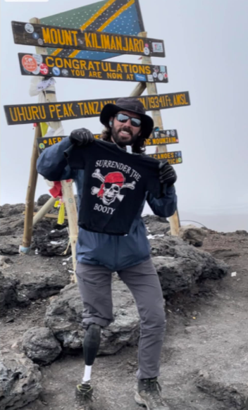 Football coach with arms crossed, standing on a football field. He wears a navy polo shirt and gray pants.