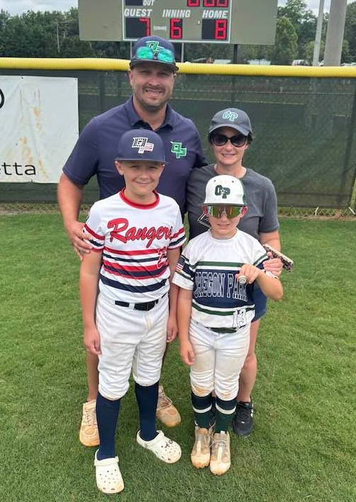 Football coach with arms crossed, looking stern on a sunny field, players in the background.