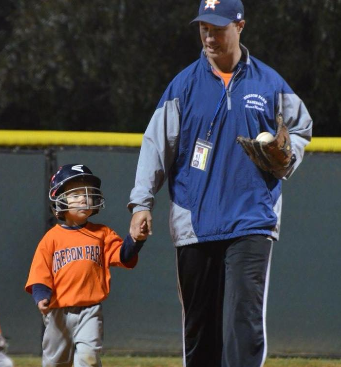 Football coach with arms crossed, looking stern on a sunny field, players in the background.