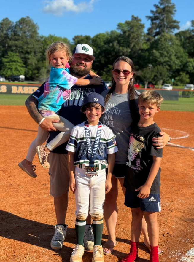 Football coach with arms crossed, looking stern on a sunny field, players in the background.