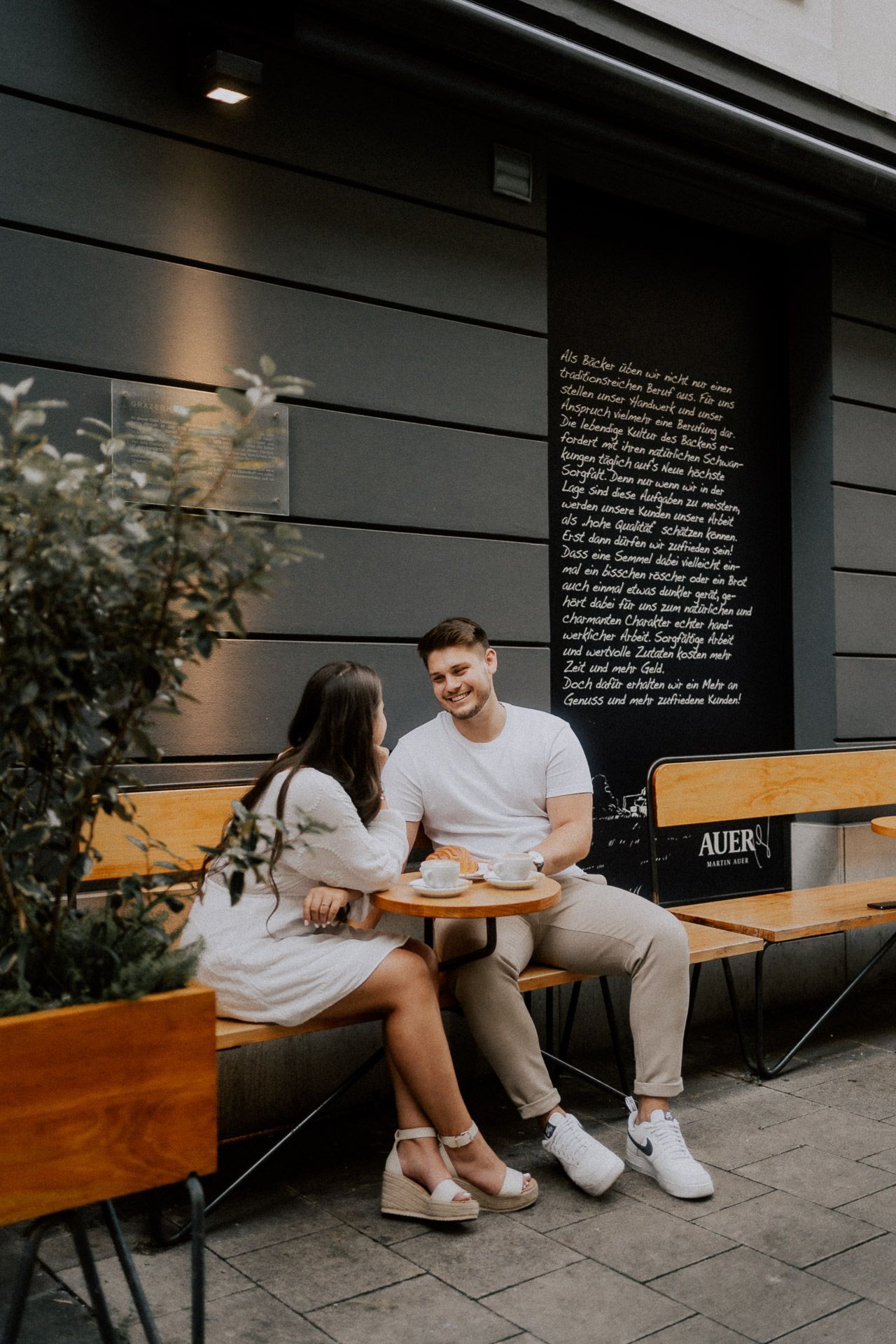 Paarfotos vor einem Café, er lacht seine Verlobte herzlich an