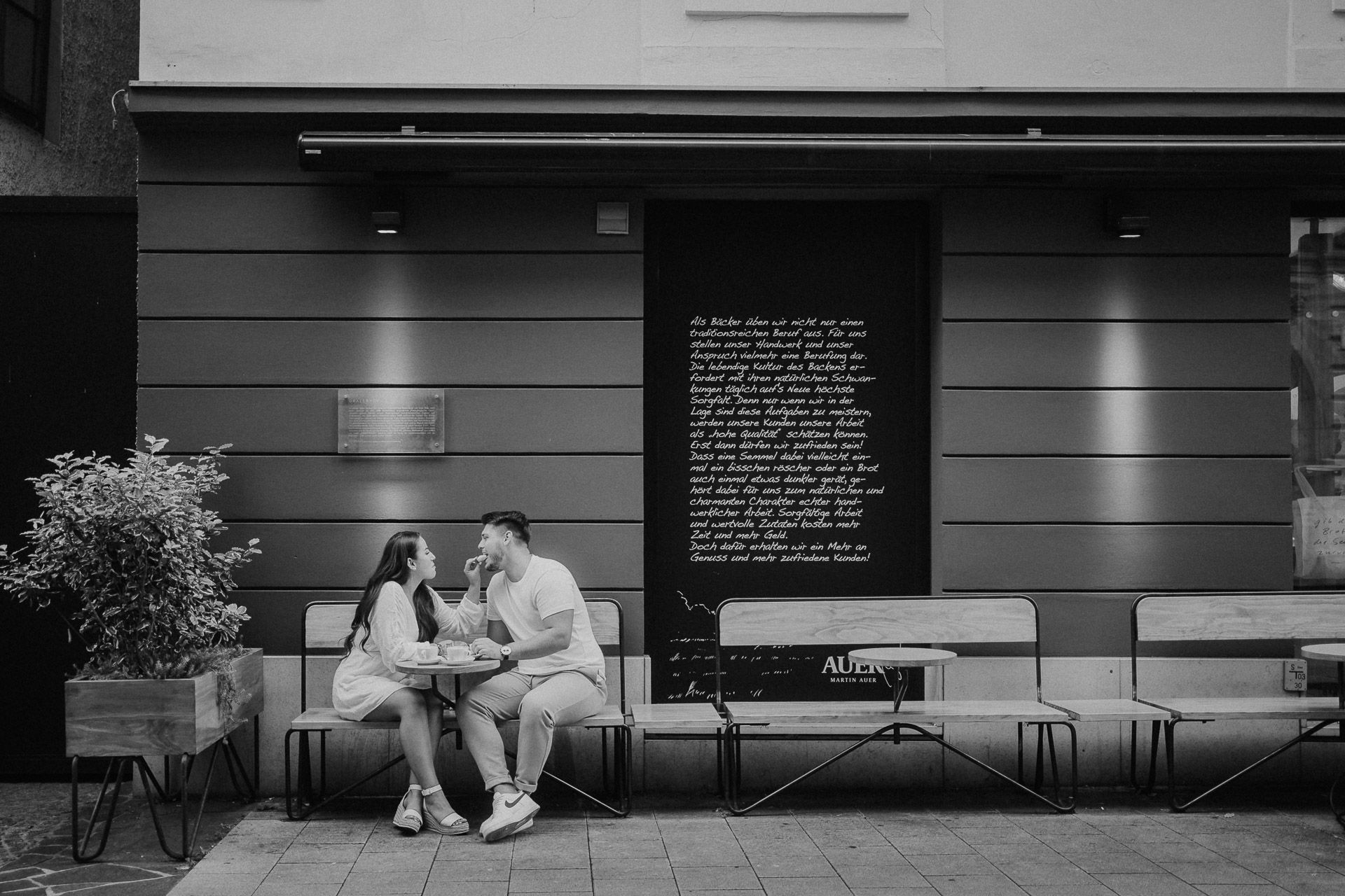 Minimalistisches Schwarz-Weiß-Bild von einem Paar, das sich vor einem Café gegenseitig Croissant füttert