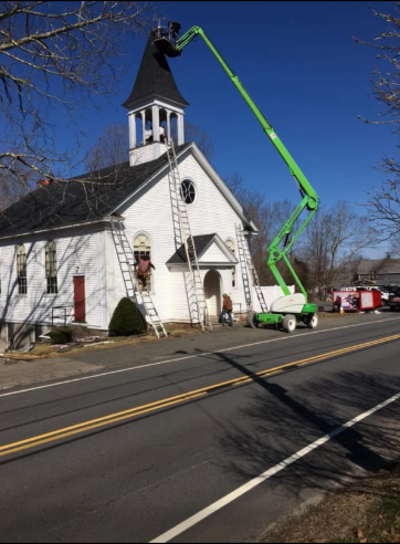Workers on ladders and a lift repairing a white church steeple on a sunny day.