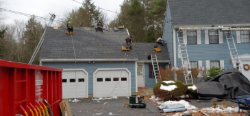 Roofers working on a house roof on a cloudy day, with multiple ladders and a red dumpster in the foreground.