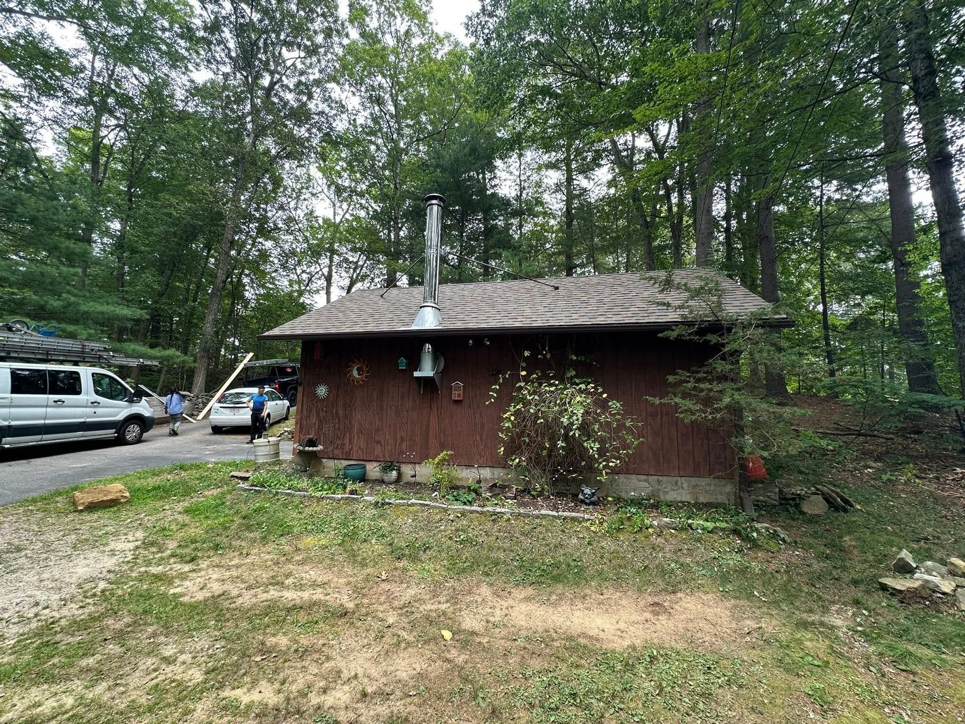 Brown cabin with a metal chimney, bell, and overgrown roof, surrounded by trees and a parked van.