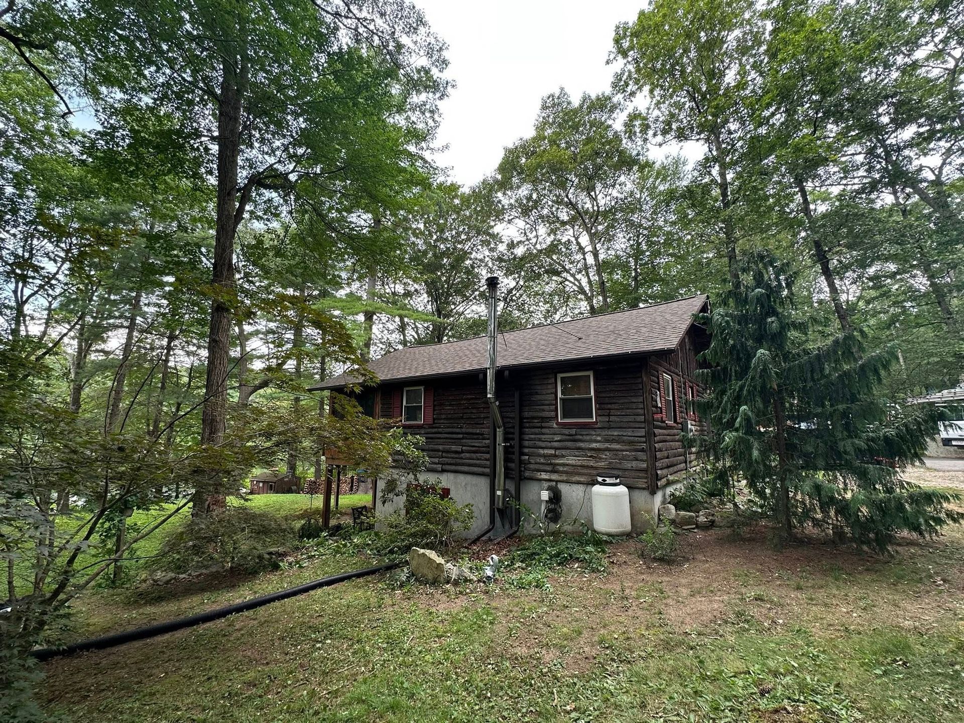 A small log cabin nestled among trees, with a chimney and propane tank visible.