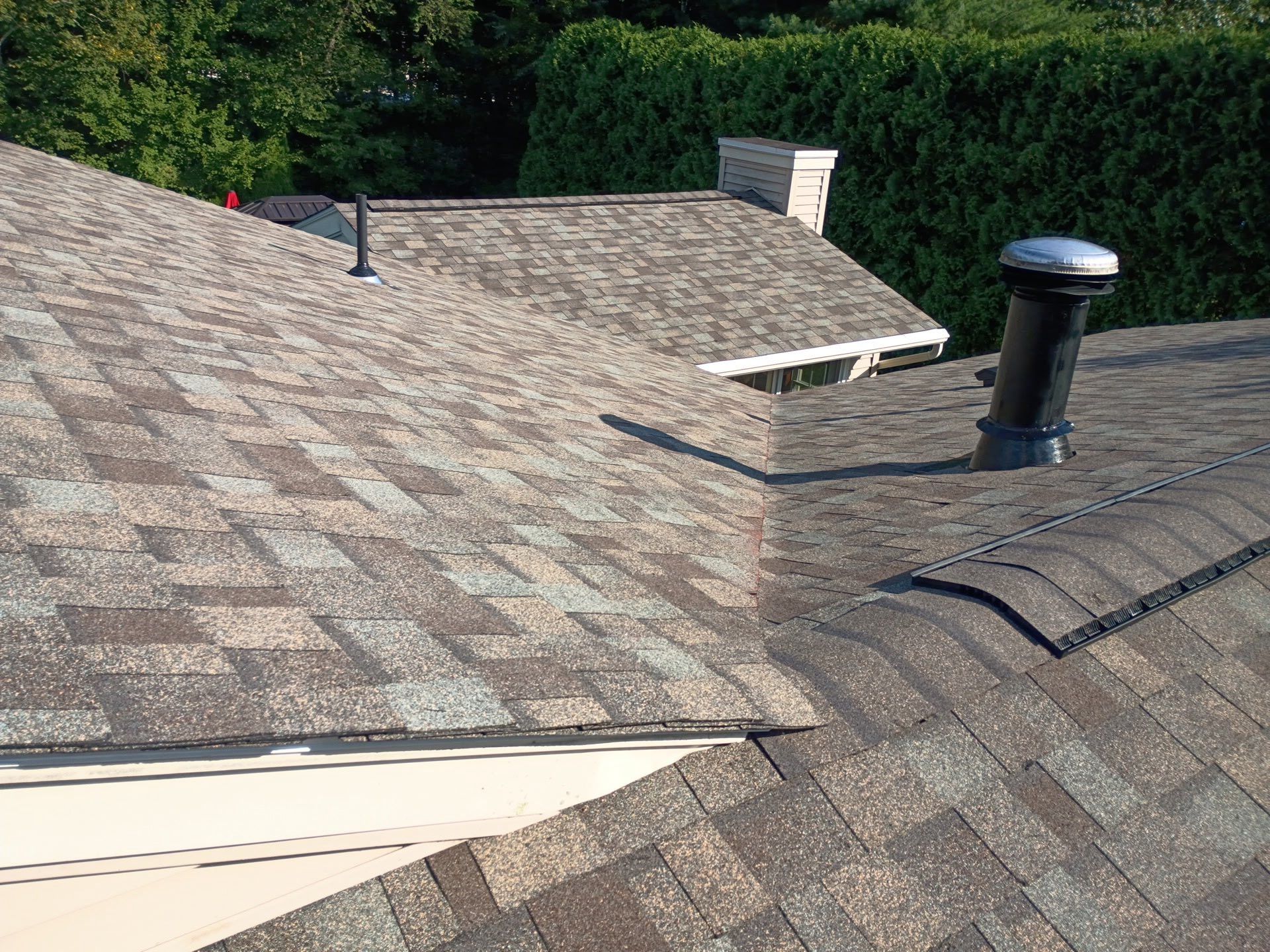 A close-up view of a shingled roof with vents, a chimney, and a natural background of trees.