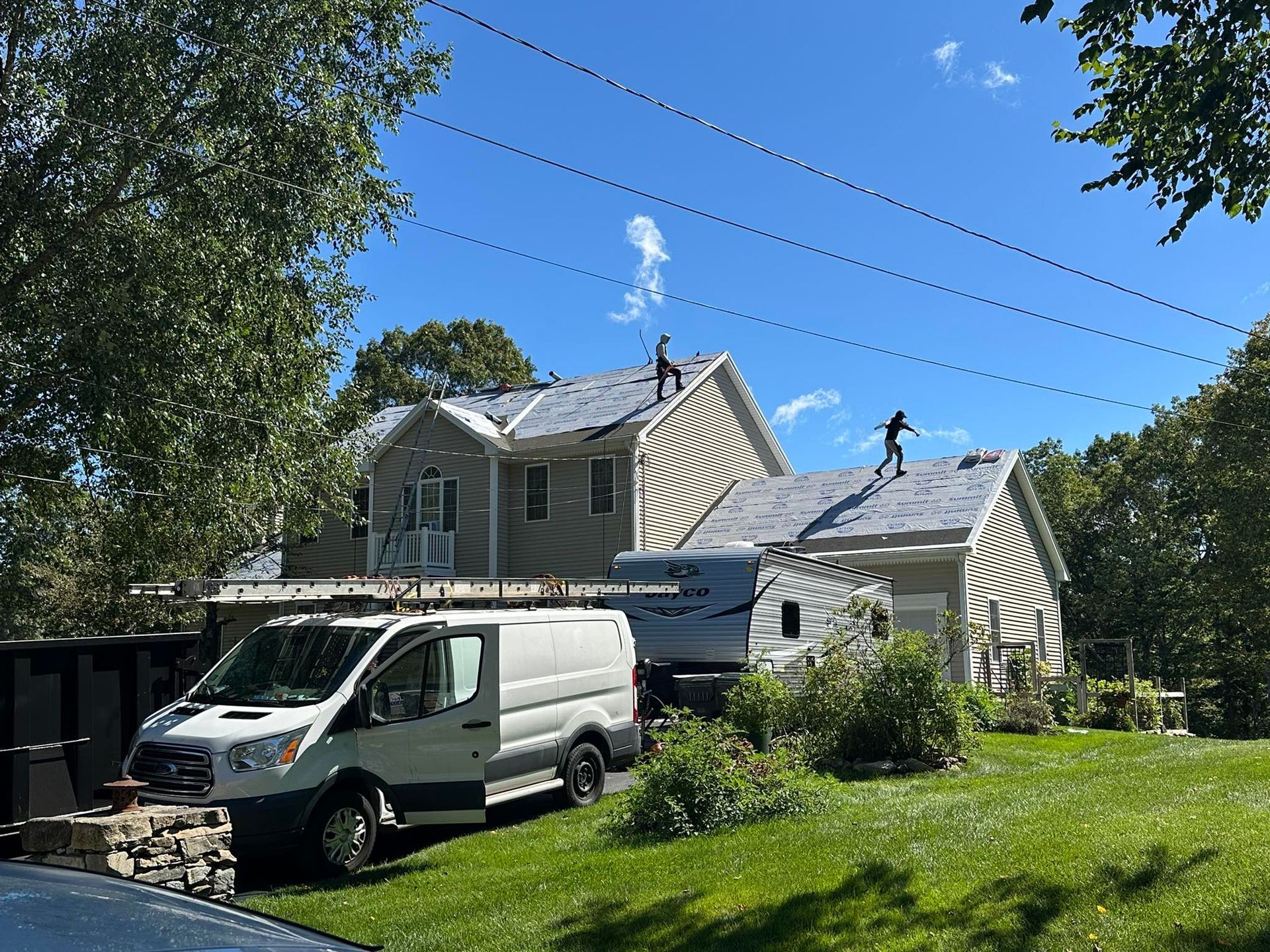View from roof: gray shingle, black ridge, ladder. Green grass and trees in the background.