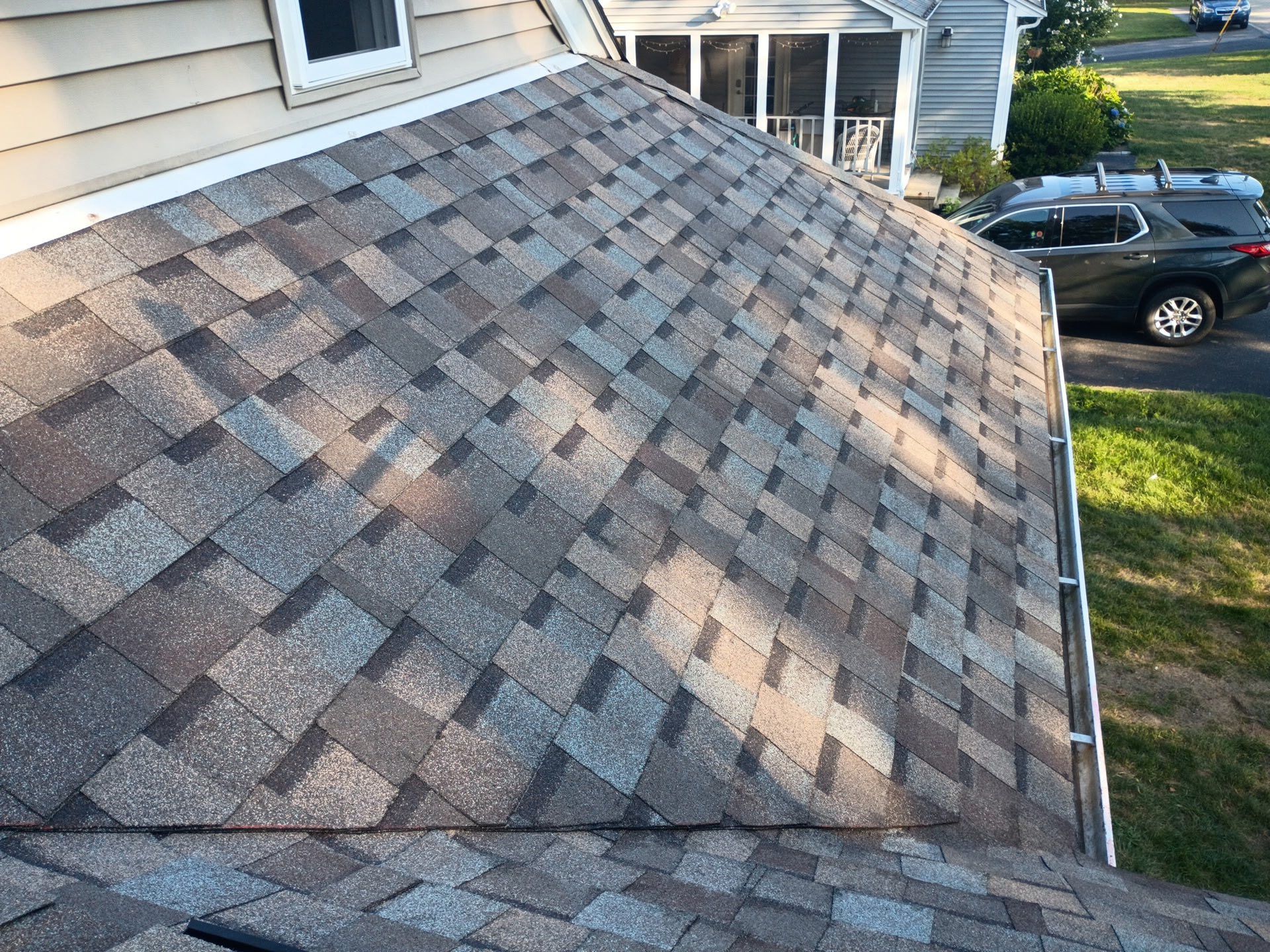 Close-up of a multi-tone gray shingle roof on a house, angled view; gutter along the edge, a car visible.