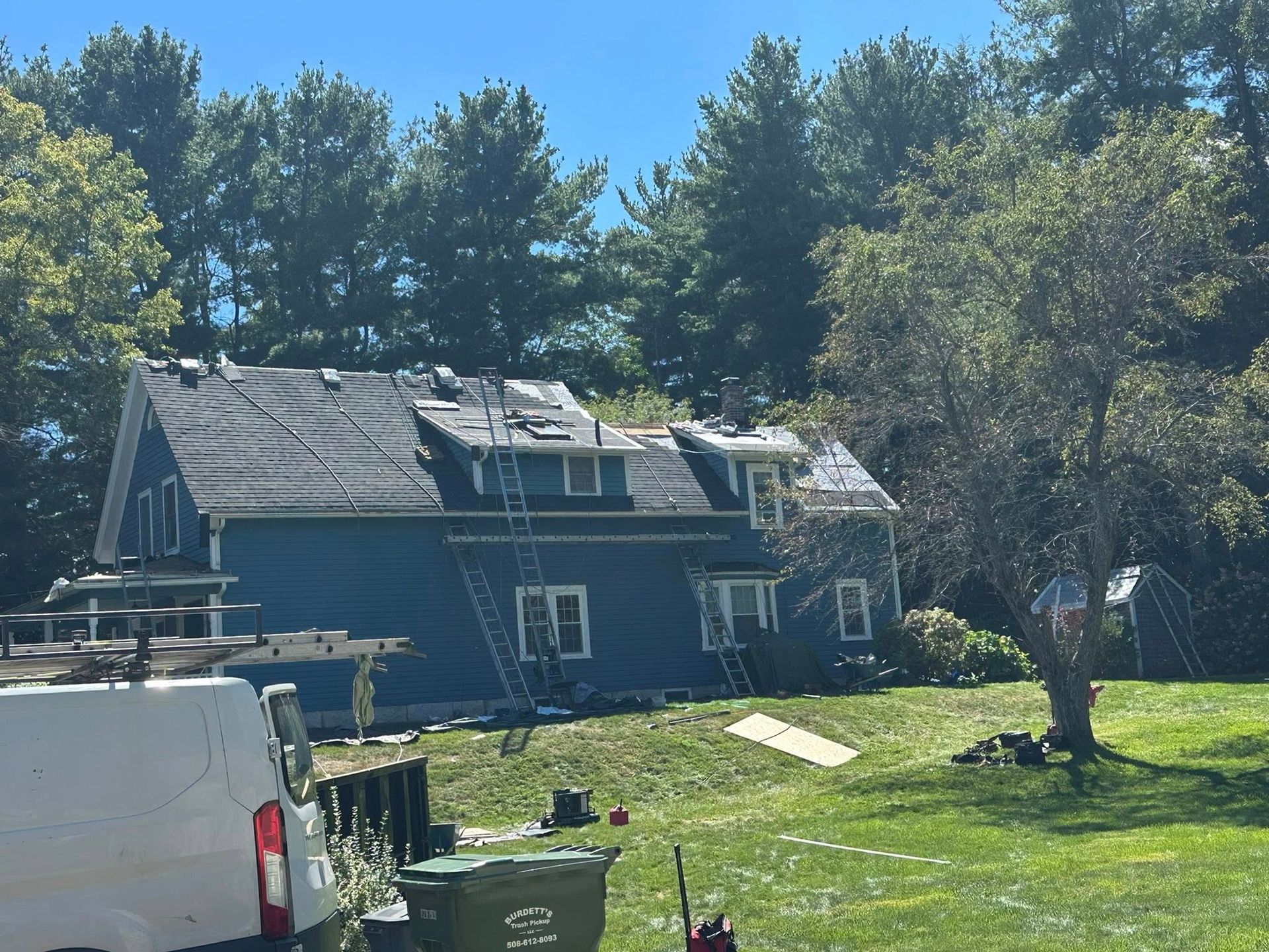 Blue house with roof replacement in progress; ladders, trees, and a van in the yard.