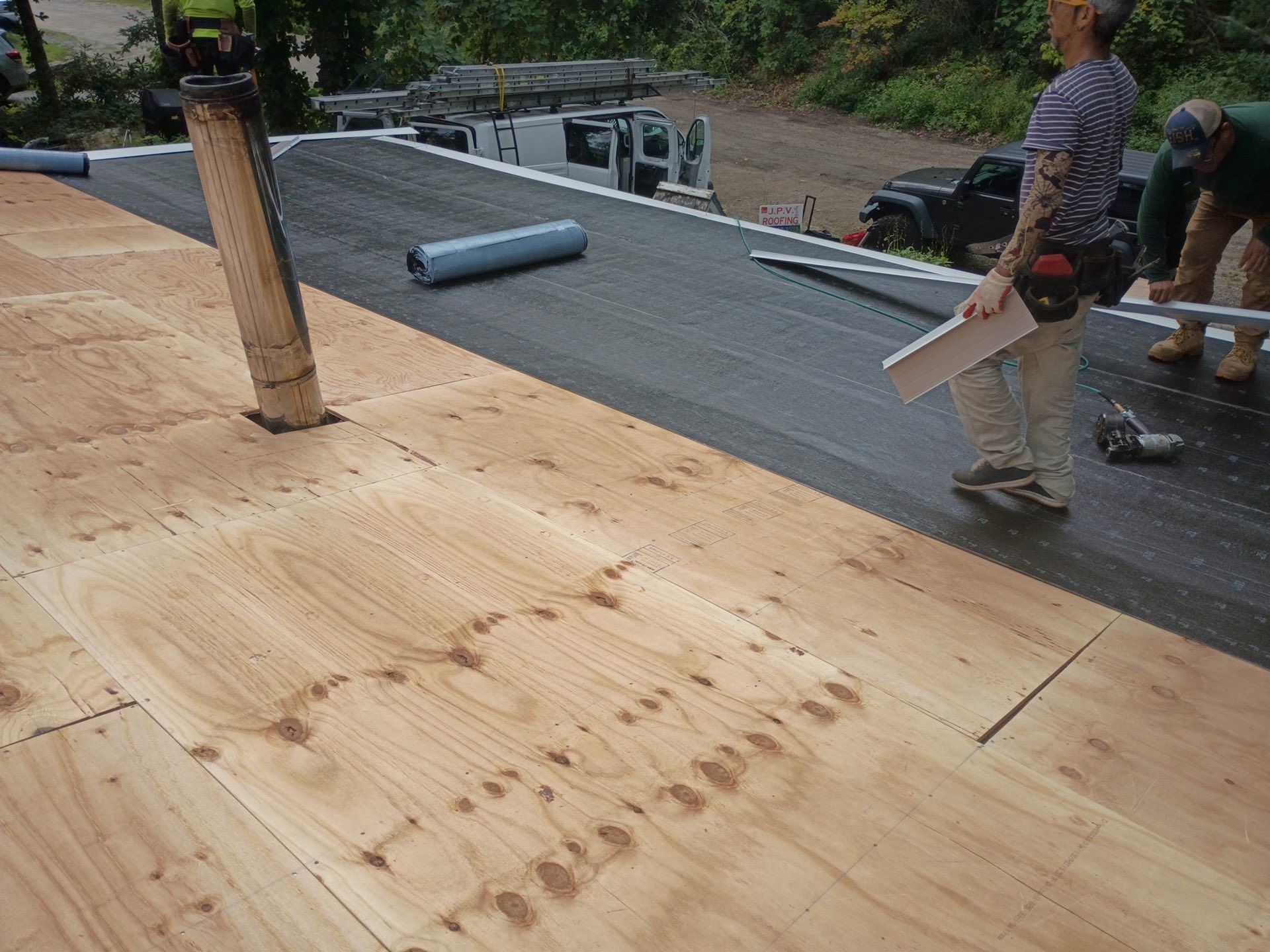 Workers installing a black roofing membrane on a wooden deck; person holding a piece of wood.