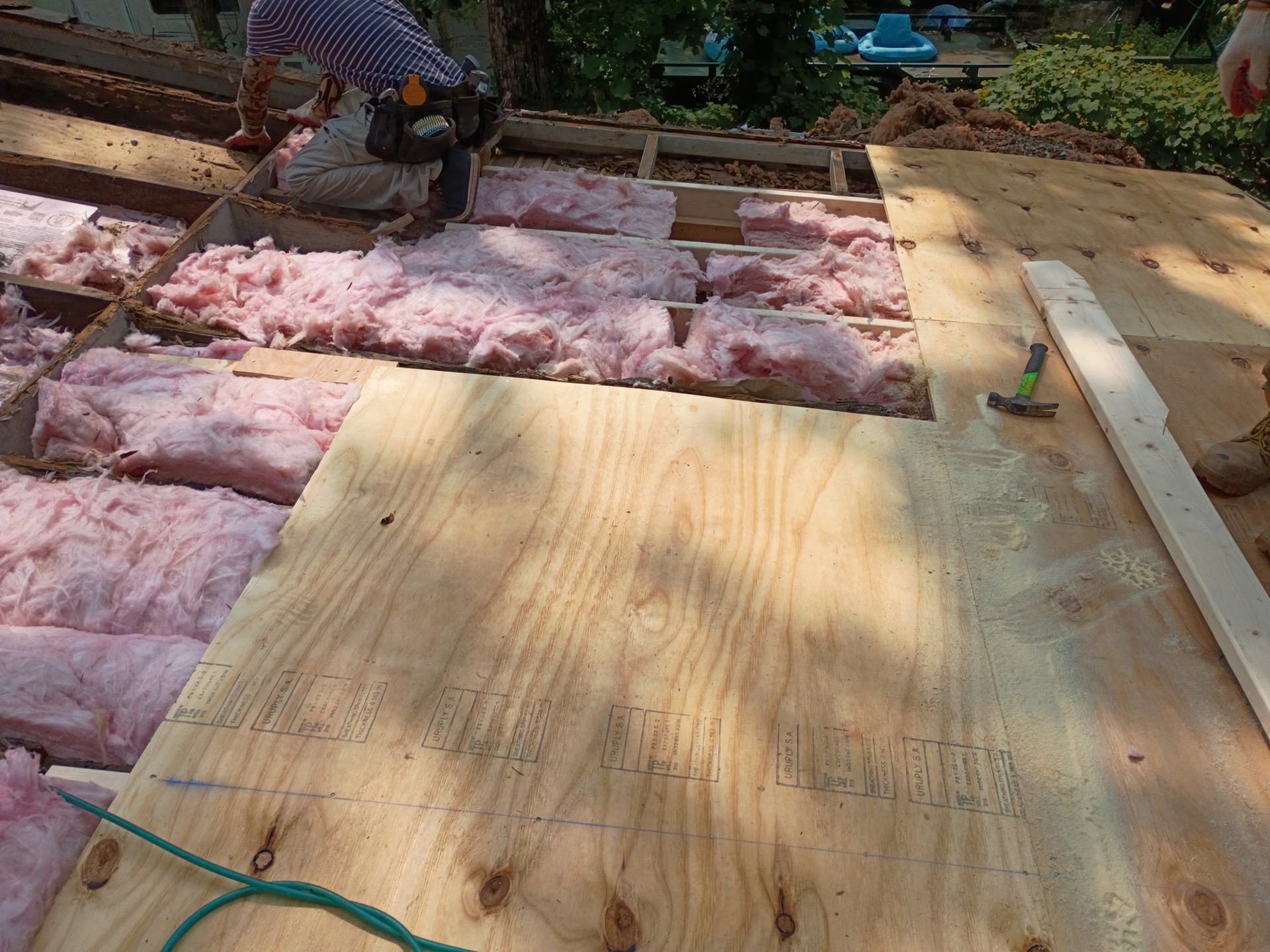 Construction worker installing plywood on roof, pink insulation visible.